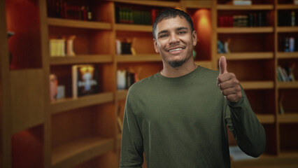 Man smiling and giving thumbs up while pointing with his index finger in front of warm wood bookshelves inside an indoor library area of a building  confident approval. © Krakenimages.com