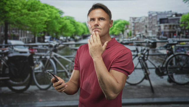 Man holding smartphone and touching chin while tapping the screen on a city street by a canal and bicycles on a bridge; contemplation decision.