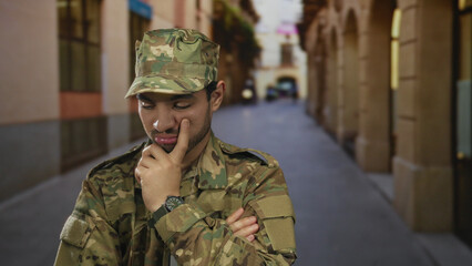 Fototapeta premium Young hispanic man in green camouflage military uniform with hand on chin contemplating on old town street; inner doubt.