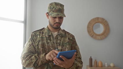 Hispanic man taps tablet in living room while wearing camouflage uniform with dog tags displayed  concentration. © Krakenimages.com