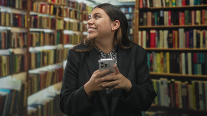Plus size hispanic woman holding a smartphone with both hands, smiling and glancing upward amid tall bookshelves in a library building  joy curiosity learning. © Krakenimages.com