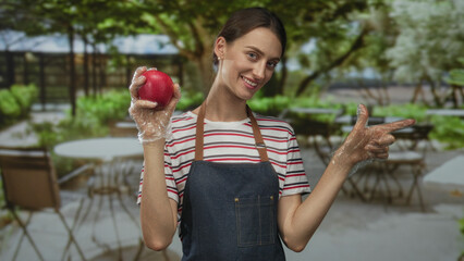 Woman holding red apple and pointing finger at outdoor cafe street terrace wearing apron and...
