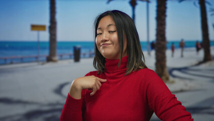 Obraz premium Young woman in red sweater pointing to herself while smiling at the seaside promenade with palm trees and ocean in the background.