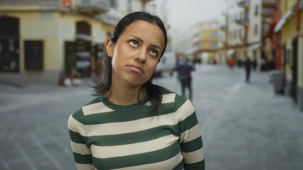 Young woman looking thoughtful on a bustling street in a cityscape, displaying emotions with her expressions, wearing a striped shirt outdoors.