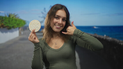 Woman holding rice cake and making call gesture on street by the seaside promenade; cheerful vacation.