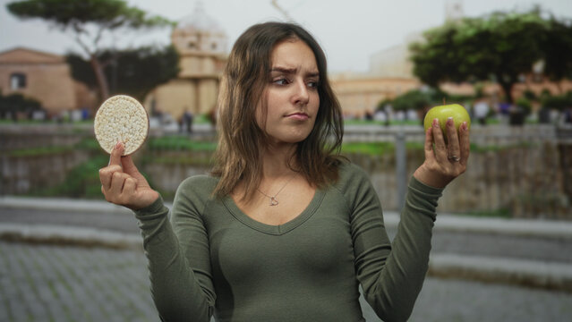 Woman holding green apple and rice cake in cobblestone street by building, holding both hands out to compare; indecision healthy choice.