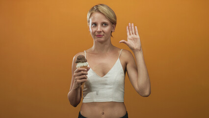Woman blond caucasian wearing cropped white top showing bare midriff holding a double scoop ice cream cone and pinching fingers gesture in studio; playful indulgence.
