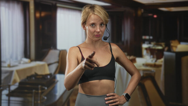 Woman in sports bra points index finger up while wearing headset in a restaurant building, hand on hip and smartwatch visible; confidence focus.