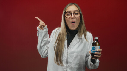 Young woman wearing white lab coat holding bottle points finger in red studio; product promotion surprise.