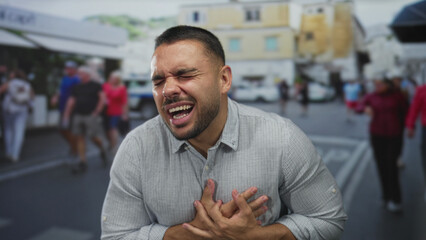 Young hispanic man clutch chest with grimace for heartattack on crowded street in bright daylight; distress.