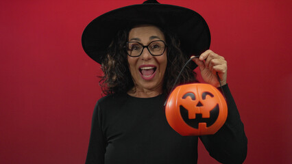 Middle age hispanic woman covers face with hand while holding a pumpkin basket in a studio; festive halloween joy surprise.