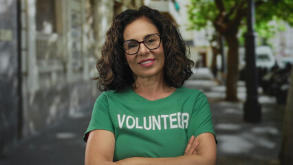 Middle age hispanic woman with crossed arms standing on a tree lined urban street wearing a volunteer uniform and glasses; denial rejection.