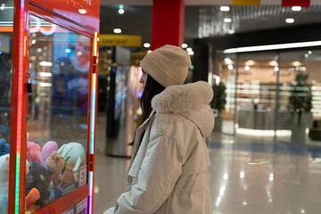 Girl stands in front of a claw machine in a busy shopping mall trying to grab plush toys
