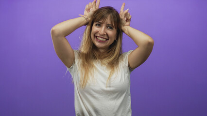 Fototapeta premium Woman smiling with hands on head making horns in a purple studio wearing a white t shirt and bracelets; playful joy lighthearted fun.