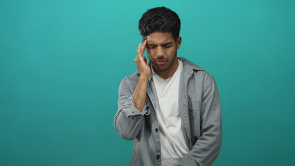 Man pressing his temple with one hand, wincing and holding his head in a teal studio with a headache; pain.