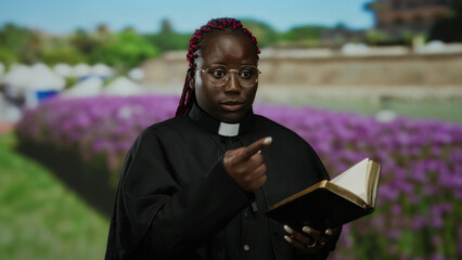 Woman reads religious book outdoors in park setting surrounded by flowers and green landscape, focusing on spirituality and serenity in a peaceful environment.