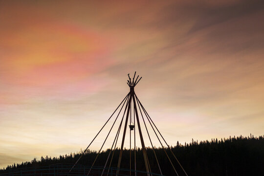View of a silhouetted teepee stands against a vibrant sunset sky, its orange and pink hues contrasting with the dark forest line, Kuusamo, North Ostrobothnia, Finland.