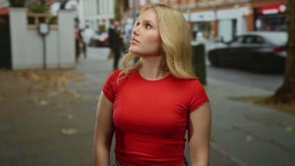 Young blonde woman in red shirt standing on a city street with blurred urban background on a sunny day, capturing a modern urban vibe with a sense of curiosity and exploration.