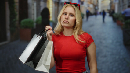 Woman shopping with bags on city street wearing red, blonde hair, casual attire, feeling tired outdoors in urban environment with people walking.