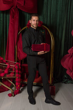 A young handsome man in a black shirt holds a gift and stands near a luggage cart in a hotel interior