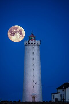 View of a full moon illuminating the stark white lighthouse tower against the deep blue sky, with a building at the base, Grottutangar, Seltjarnarnesbaer, Iceland.