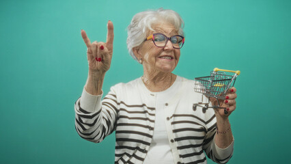 Woman wearing striped cardigan and eyeglasses holding a miniature shopping cart and flashing rock hand sign in studio  playful joy. © Krakenimages.com
