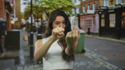 Fototapeta premium Hispanic woman in white tank top with clenched fist, extended middle finger and furrowed brow on street; anger.