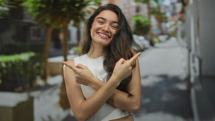 Fototapeta premium Young woman pointing finger toward opposite directions on a sunlit street corner lined with potted plants; guidance direction clarity optimism.