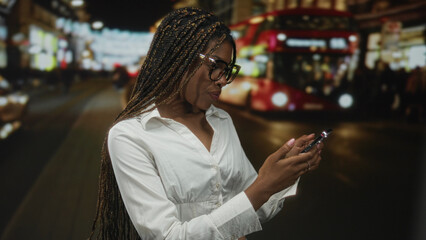 Woman wearing glasses with long braids smiling and typing on smartphone while standing on a busy city street at night; urban connection joy.