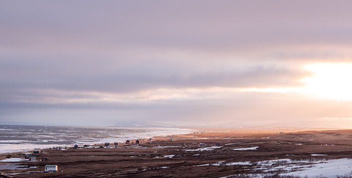 View of a tundra landscape meeting the ocean under a pastel sky during sunset, with scattered buildings and patches of snow, Nome, Alaska, United States.
