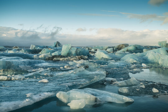 View of shimmering icebergs with crystal blue hues float serenely on the glacial lagoon under a soft, cloudy sky, Jokulsarlon, Sveitarfelagid Hornafjordur, Iceland.