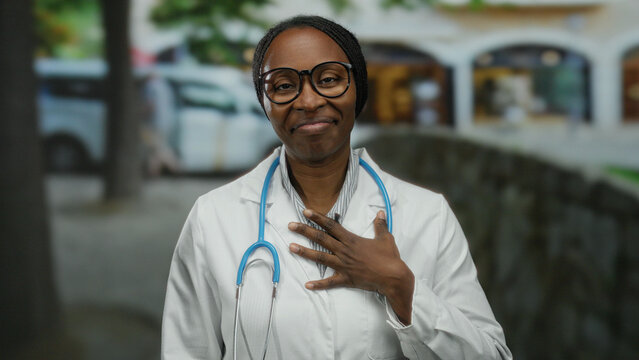 Woman outdoors smiling with hand on chest wearing glasses with stethoscope on street in urban cityscape background.