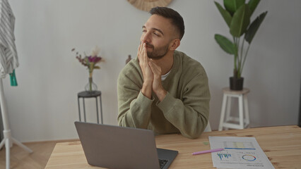 Young man sitting at table with laptop in living room looking thoughtful while working indoors