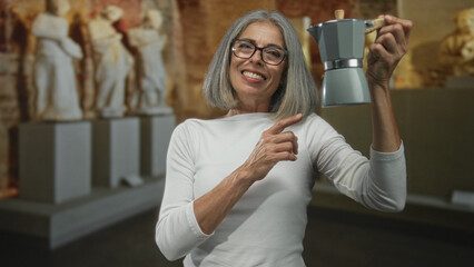 Woman points finger to moka pot while holding it in a building gallery surrounded by sculptures;...