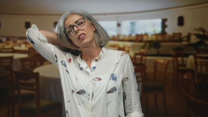 Woman holding neck with hand in restaurant building, wearing glasses and floral blouse; visible discomfort.