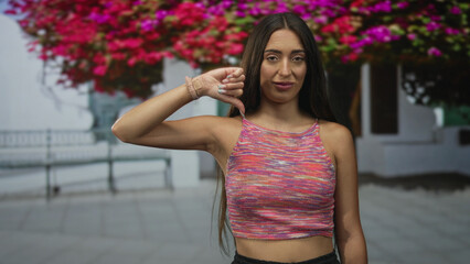 Young hispanic woman showing thumbs down with exposed thumb and hand in street by white building,...
