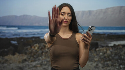 Young hispanic woman holding smartphone and looking at screen on rocky pebble beach shore in casual tanktop; calm reflection.