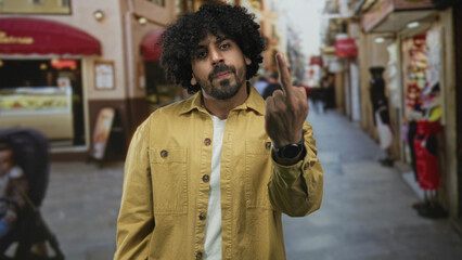Man shows middle finger with both hands on a crowded street, curly hair and yellow jacket visible;...