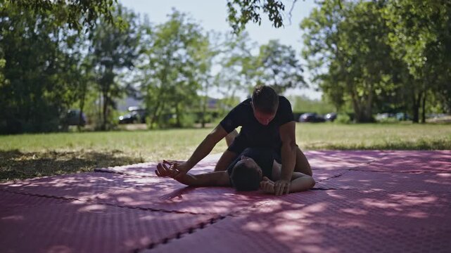 Two young boys engage in intense mixed martial arts wrestling practice on a red mat in a sunny park. They demonstrate grappling techniques and discipline during their outdoor training session.