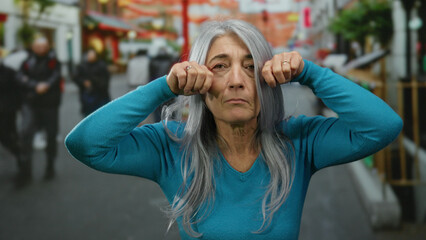 Fototapeta premium Senior grey-haired woman in blue sweater playfully gesturing on a busy outdoor street with blurred pedestrians in the background, capturing a candid urban scene full of life.