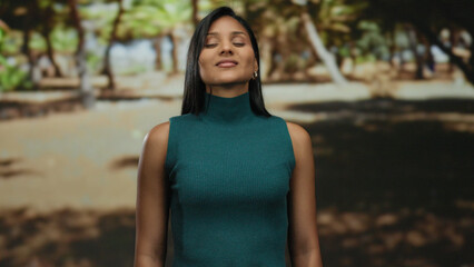 Young hispanic woman relaxing outdoors in a serene park, standing amidst lush greenery and sunlight, exuding calm and tranquility, eyes closed in peaceful meditation.