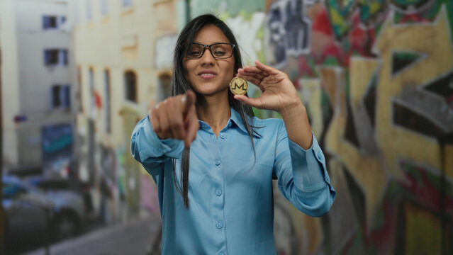 Woman smiling while holding a monero coin and pointing at the camera, standing outdoors against a colorful urban street backdrop, symbolizing digital currency enthusiasm.