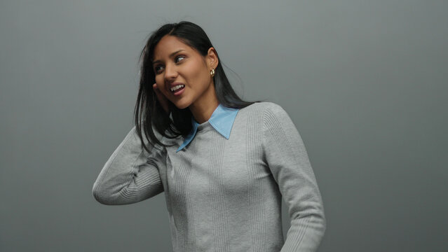 Young hispanic woman makes a listening gesture with her hands on ears, standing against a grey background, conveying curiosity and attention in a casual setting.