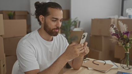 Man holding smartphone with both hands at table in building surrounded by moving boxes and vase; quiet concentration.