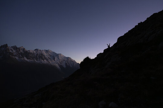 View of a small figure standing on a dark slope with hands up, silhouetted against the backdrop of snowy mountains and a gradient sky, Chamonix-Mont-Blanc, Auvergne-Rhone-Alpes, France.