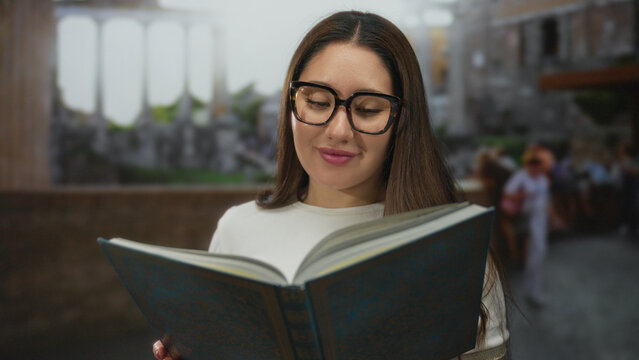 Young hispanic woman reading large book and smiling outdoors at roman ruins with oversized glasses and long hair; serenity knowledge culture exploration.