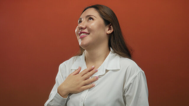 Young hispanic woman in white shirt holds hair in studio with deep red walls; confidence selfcare empowerment grace.