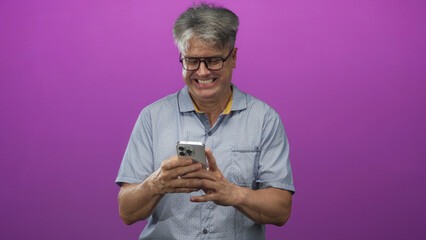Fototapeta premium Man with grey hair and glasses holding smartphone, tapping screen with hands while smiling in studio; joy connection.