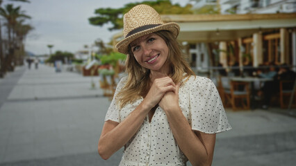 Woman points both index fingers to camera on street terrace restaurant wearing straw hat and polka dot dress; playful affection.