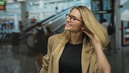 Woman smiling in airport terminal indoor with blonde hair and glasses, capturing a candid moment in busy environment with escalator background and passengers.
